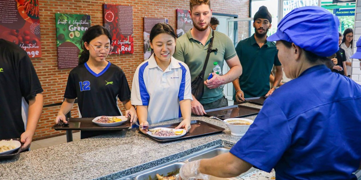 Happy students queue for healthy international school lunch while staff serves food in a clean canteen