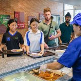 Happy students queue for healthy international school lunch while staff serves food in a clean canteen