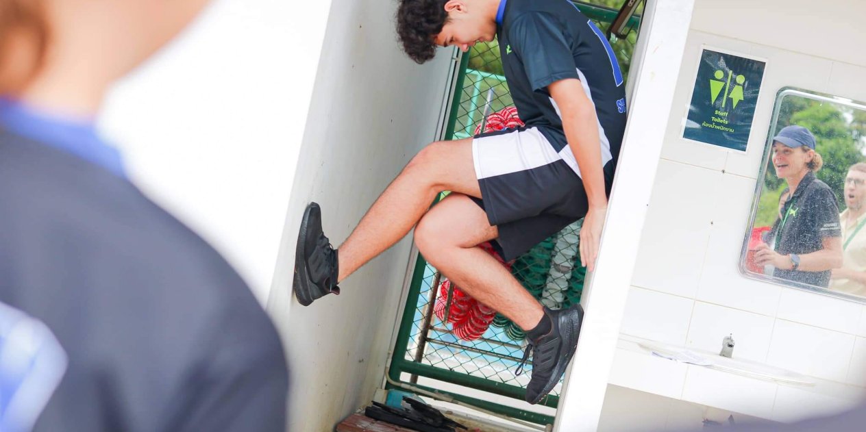 Student climbing during a school physical challenge while teachers cheer supportively, encouraging confidence and exploration in sport